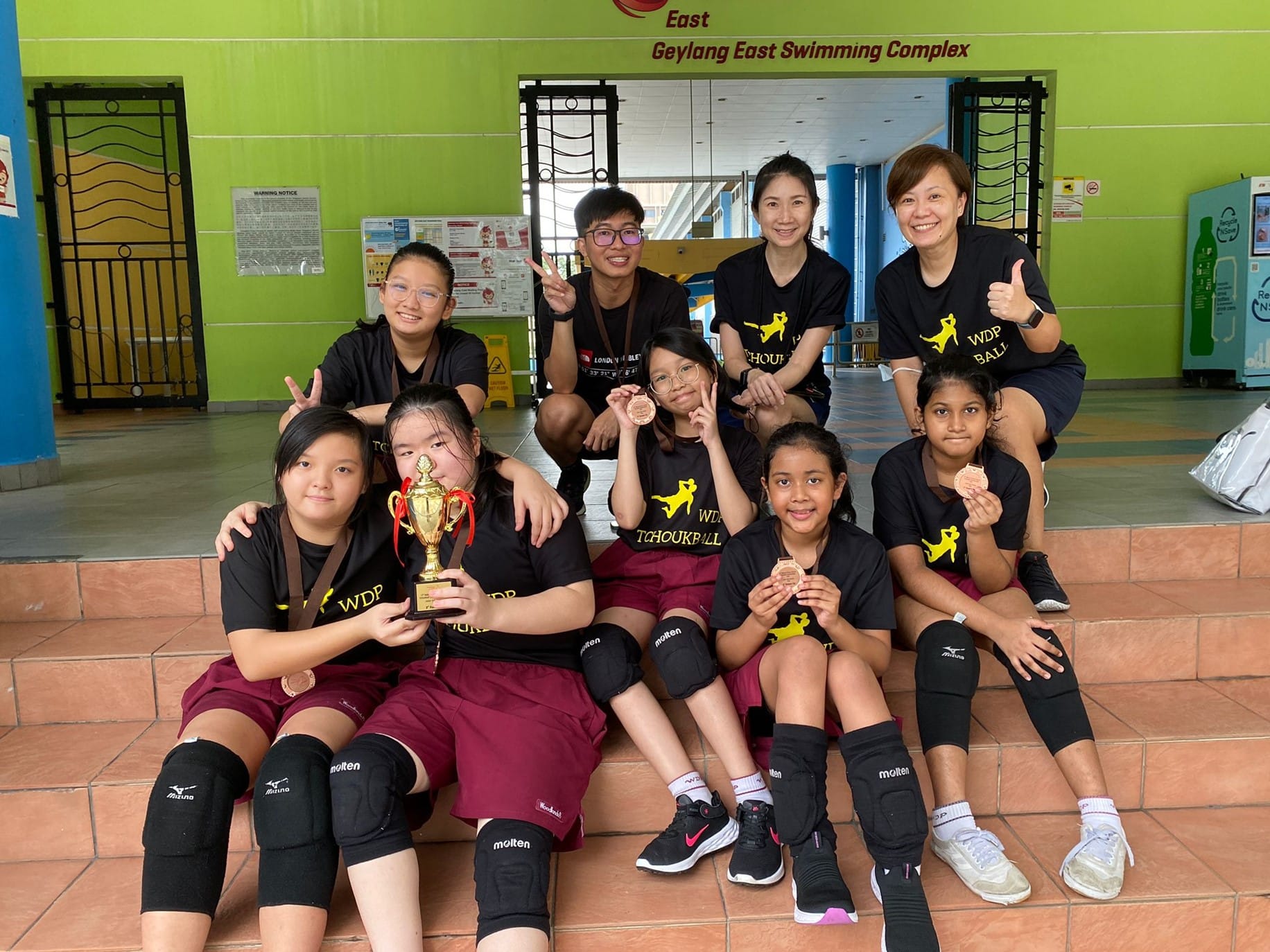A youth tchoukball team in black WDP shirts poses with a trophy and bronze medals at Geylang East Swimming Complex.